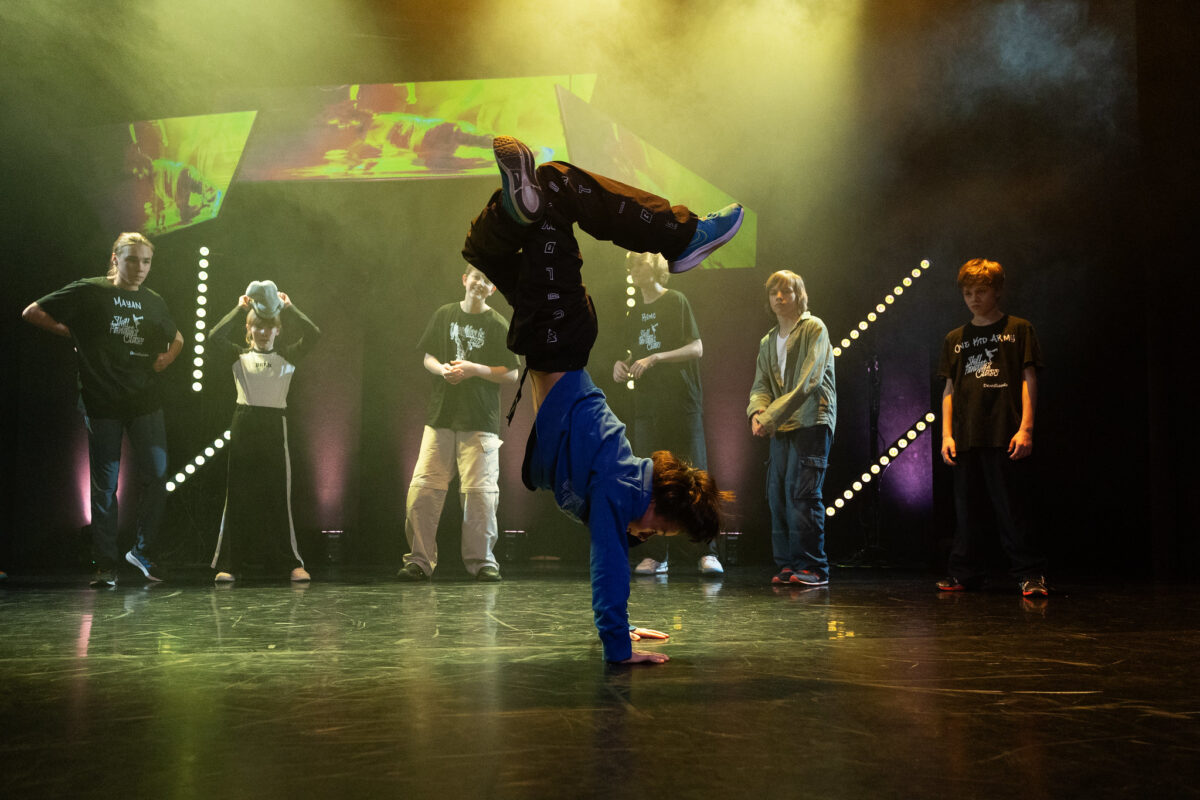 Chłopak tańczy breakdance na scenie, stoi na rękach, za nim grupa nastolatków