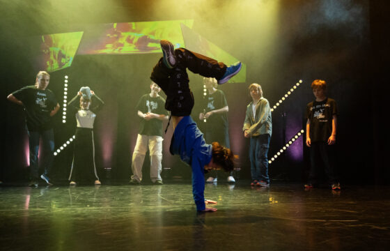 Chłopak tańczy breakdance na scenie, stoi na rękach, za nim grupa nastolatków