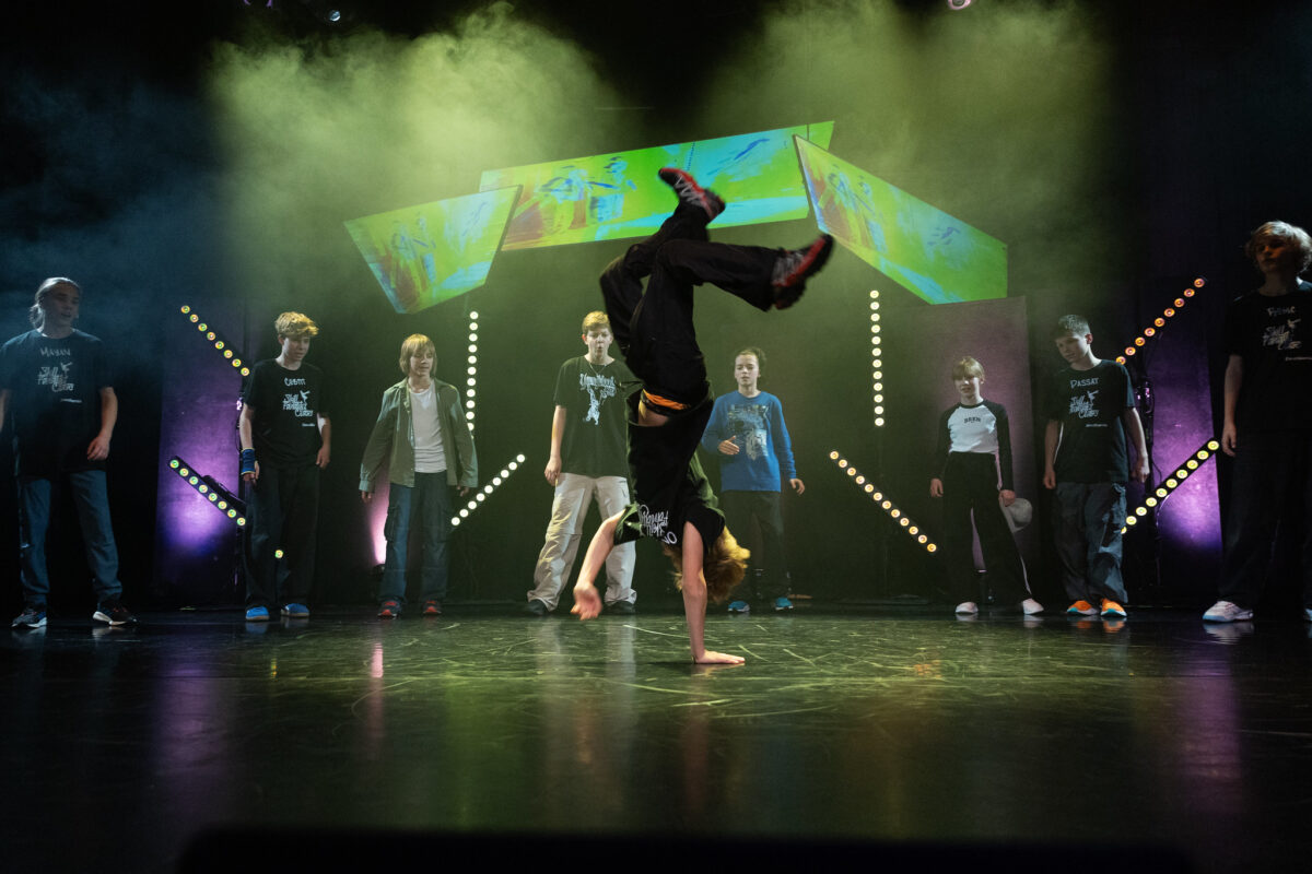 Chłopak na scenie tańczy breakdance, stoi na jednej ręce, zanim grupa dzieciaków