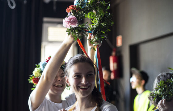 Na pierwszym planie uśmiechnięta dziewczyna, za nią druga próbuje założyć jej na głowę wianek z żywych kwiatów