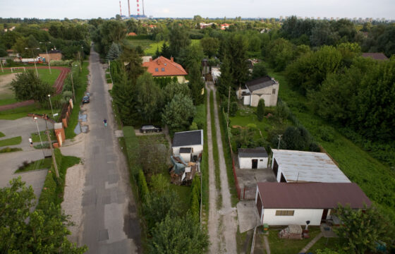 Wąska asfaltowa droga, po jej lewej stronie boisko, po prawej budynki. Po obu stronach drogi drzewa. Zdjęcie zrobione z góry