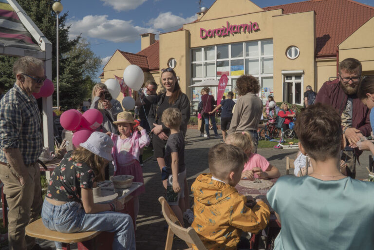 Grupa ludzi, dorosłych z dziećmi na podwórku przed budynkiem z napisem Dorożkarnia, są prawdopodobnie na pikniku