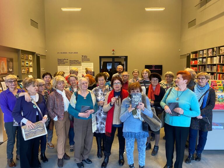 Duża grupa kobiet - seniorek, pozuje do zdjęcia prawdopodobnie w jakiejś bibliotece, księgarni lub galerii.