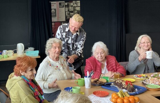 Cztery starsze kobiety siedzą przy zastawionym stole, uśmiechnięty mężczyzna za nimi trzyma mikrofon przed jeną z nich