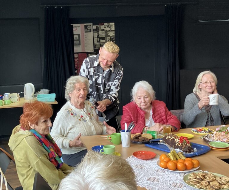 Cztery starsze kobiety siedzą przy zastawionym stole, uśmiechnięty mężczyzna za nimi trzyma mikrofon przed jeną z nich