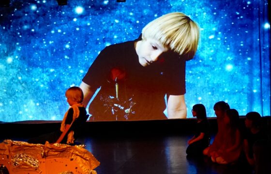 Scena teatralna, na dużym ekranie postać chłopca, przed ekranem ten sam chłopiec i grupa dzieci