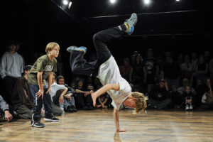 Chłopak na parkiecie tańczy breakdance, wykonuje figurę stojąc na jednej ręce, wokół siedzą dzieci i młodzież