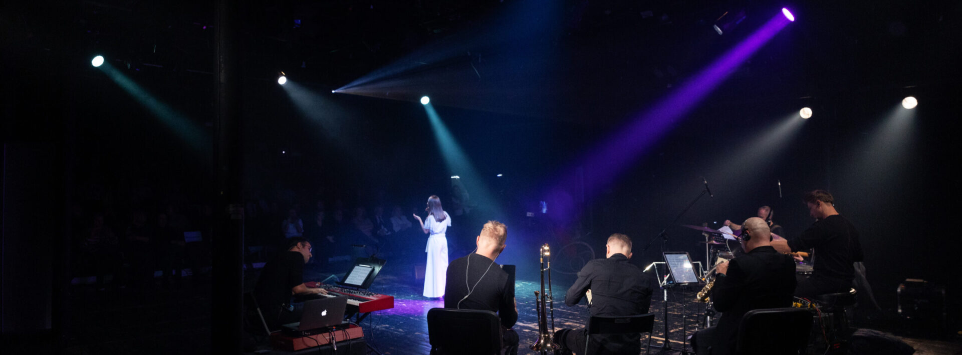 Grupa muzyków i postać w długiej suknie na scenie przed widownią. Zdjęcie zrobione na widownię.